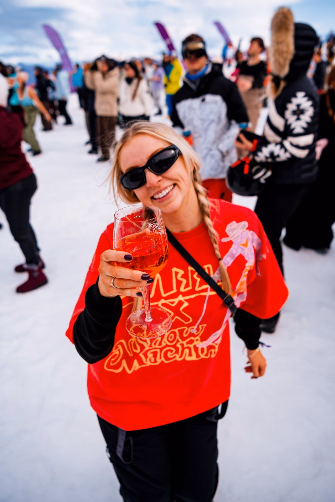 Woman with a side braid and oversized sunglasses holding up an Aperol Spritz on the mountain